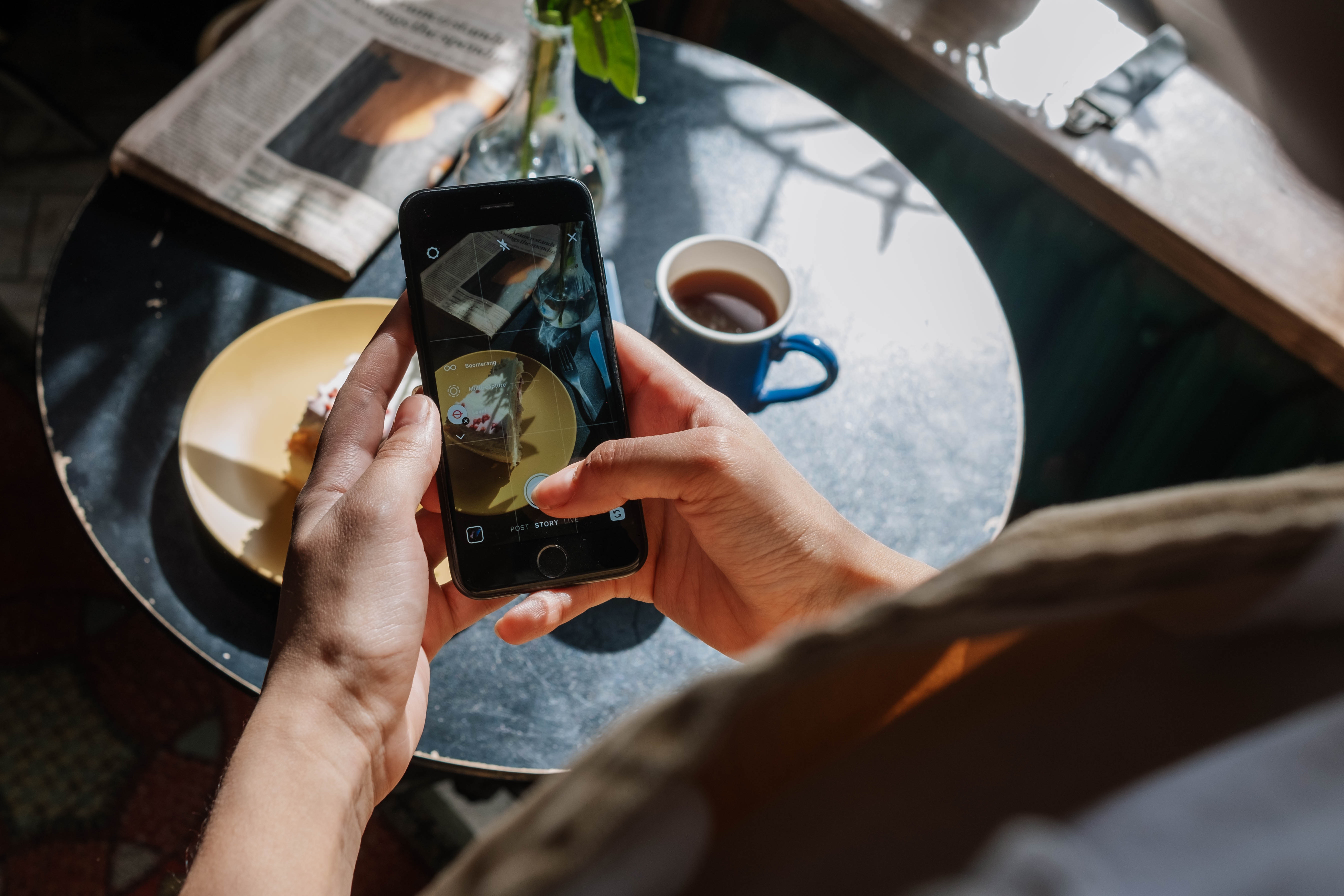 Person holding their phone and taking a photo of their newspaper, pastry and coffee.